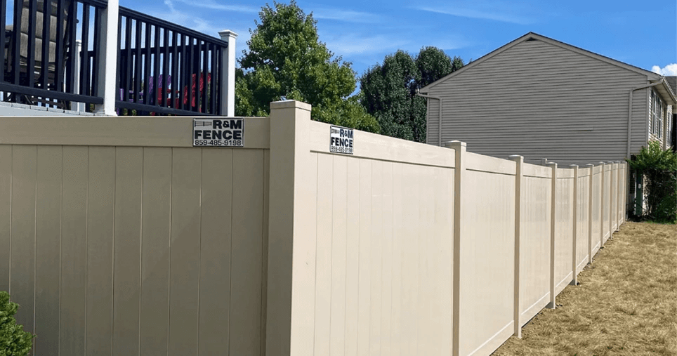 Tan vinyl privacy fence running along a sloped backyard beside a home.